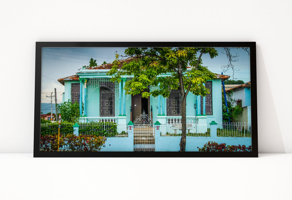 Poster Cuba Villa 3 ingelijst, kleurenfoto van een lichtblauwe villa achter een groene boom, met smeedijzeren ramen en een tuin vol bloemen